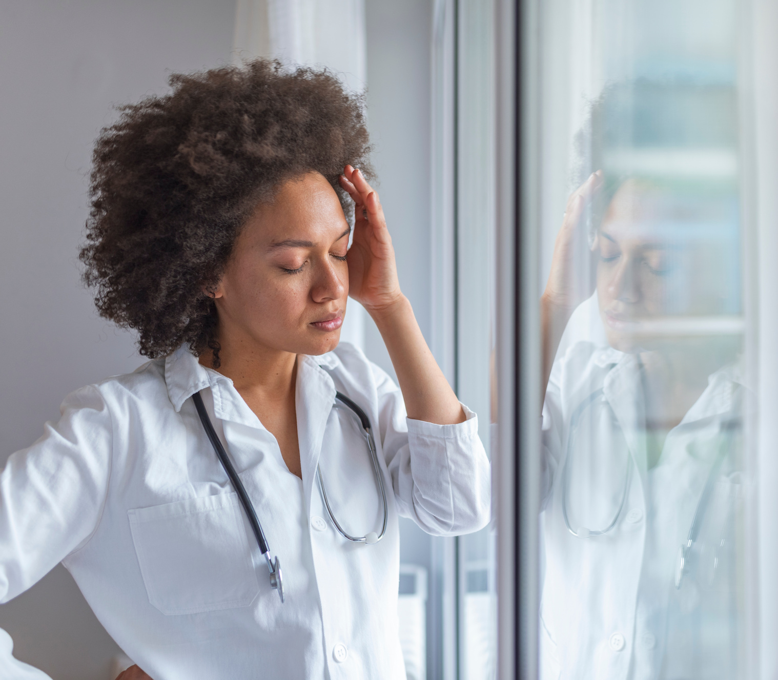 Sad female doctor leaning against the wall in hospital corridor. Female doctor with head pain standing near window. It's a stressful profession. Upset female nurse standing in hospital corridor